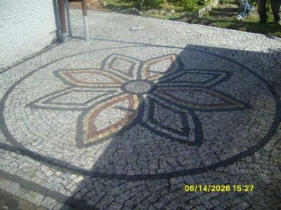 Okrągły wzór kwiatowy ułożony z kostki granitowej w odcieniach szarości, brązu i czerni, otoczony szarą kostką, widoczny cień rzucany przez budynek.